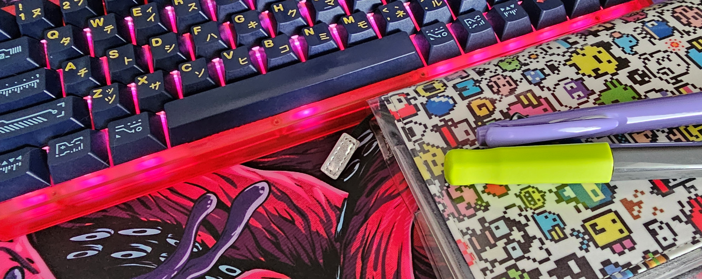 Pink and grey keyboard and planner with a pixel monster cover. A green and a purple pen are on the planner.